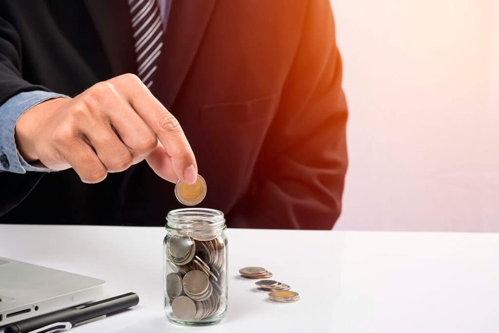Grupo Albatros. Mano de señor con moneda de 2€ haciendo el gesto de introducirla en un tarro de cristal con más monedas. El tarro se encuentra sobre una mesa blanca junto a monedas de euro.