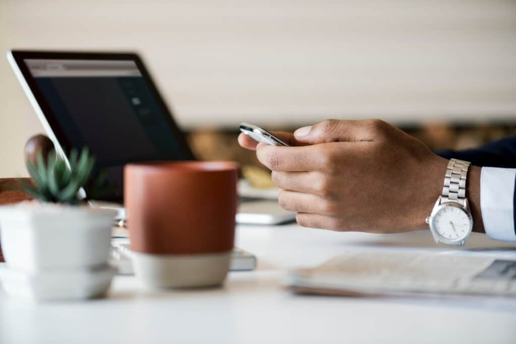 manos de persona con teléfono móvil entre ellas, ordenador portátil al frente y vaso de café al lateral