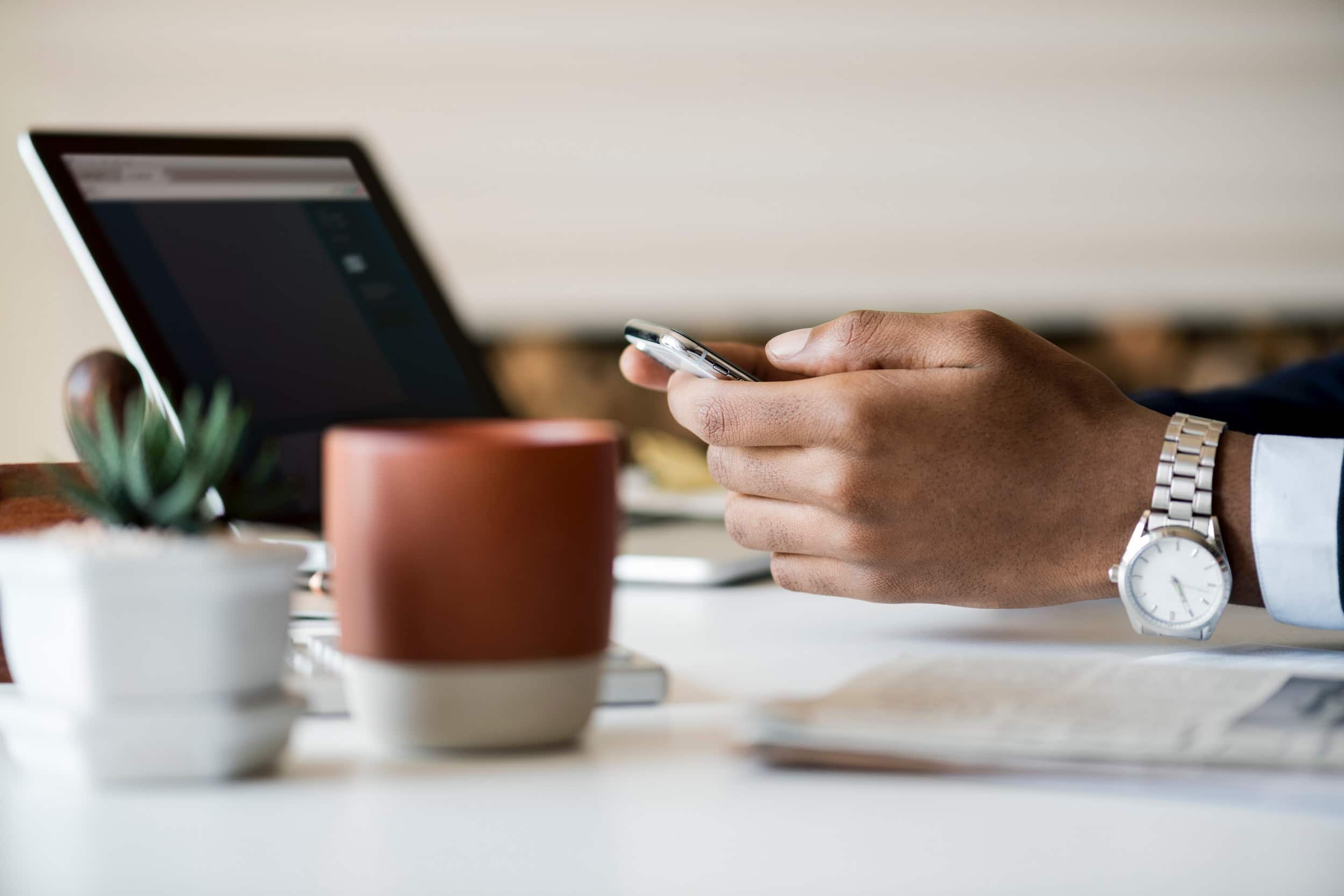 manos de persona con teléfono móvil entre ellas, ordenador portátil al frente y vaso de café al lateral